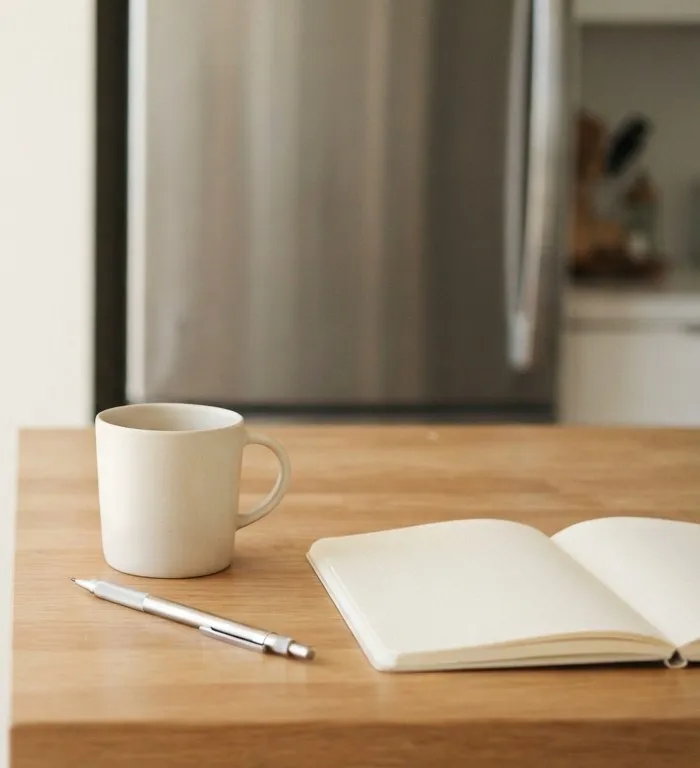 Coffee mug and open notebook on a kitchen table near a refrigerator